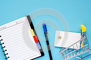 Mini shopping cart or trolley with blank credit chip card, spiral notebook and pen on blue background