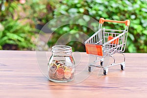 Mini Shopping Cart or Trolley and Glass Jar Filled with Coins Isolated on a Table