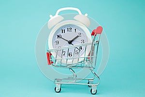 Mini shopping cart with white alarm clock on blue background. Conceptual image of sale, seasonal discounts in shopping