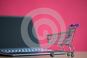 Mini shopping trolley stands in front of a laptop on a pink background. Online shopping concept. Black computer screen