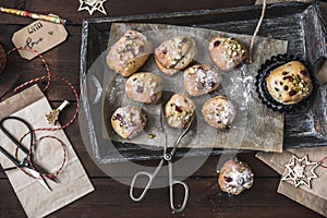 Mini stollen with sugar icing on a wooden tray, paper bags, scissors, pen holder and gift ribbon on dark wood