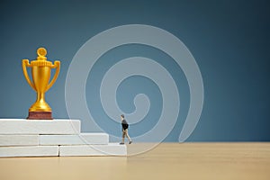 Miniature business concept - businessman walking on white staircase ladder to reach golden trophy