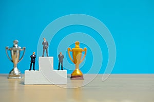 Miniature business concept - three businessman standing on winner podium with golden and silver trophy