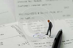 Miniature business man figurine standing on printed payment invoice, bill or receipt and looking at VAT with pen circle using as photo