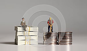 A miniature businessman sitting on a pile of $100 bills and a miniature worker on a pile of coins.