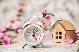 Miniature clock and small wooden house close-up and copy space. Alarm clock, house, flowering branch in the spring. The concept of