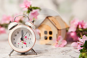 Miniature clock and small wooden house close-up and copy space. Alarm clock, house, flowering branch in the spring. The concept of