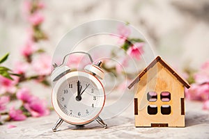 Miniature clock and small wooden house close-up and copy space. Alarm clock, house, flowering branch in the spring. The concept of