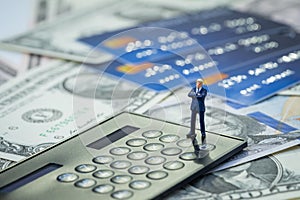 Miniature confidence businessman standing and thinking on black calculator on pile of credit cards and US Dollar bills money using