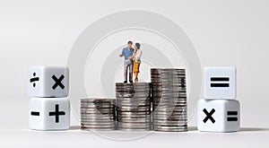 A miniature couple standing on a pile of coins with arithmetic symbols cube.