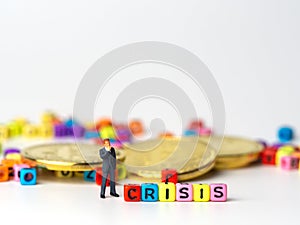 Miniature figure businessman in dark blue suit standing backside of colorful of CRISIS alphabet and golden coin and thinking of th