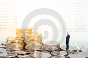 Miniature figure businessman stand on pile of coins thinking and looking to stack of coins with bank passbook as background.