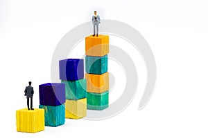 Miniature figurine posed as businessman sitting alone on a wooden cube looking up to leader on colourful stacks as business chart