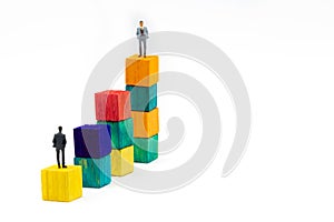 Miniature figurine posed as businessman sitting alone on a wooden cube looking up to leader on colourful stacks as business chart