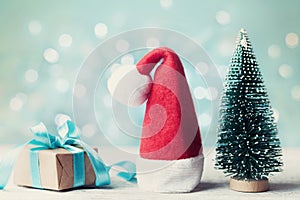 Miniature fir tree, santa hat and christmas gift box against blue bokeh background. Holiday greeting card.