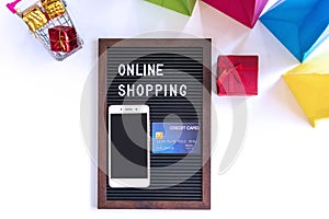 Miniature of gift boxes in trolley, smartphone, word on black frame, credit card and colorful bags on white desk. Online shopping