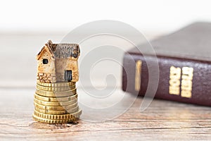 Miniature house on top of a golden stack of coins with a closed Holy Bible Book on a wooden table