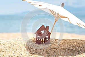 Miniature house and umbrella on beach, blue sea, sky on background. Real estate, property investment concept. Copy space