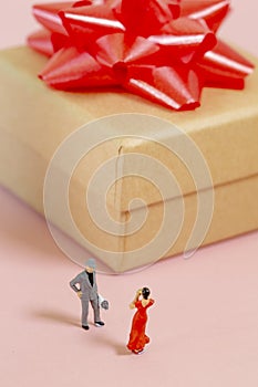 A miniature man standing in front of a woman with flowers in his hands and a big gift box behind him for valentines day, birthday