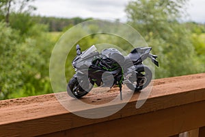 A miniature model of a black sports motorcycle - perched on a wooden railing - overlooks a lush green landscape - cloudy sky