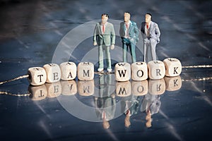 Miniature model of businessmen and wooden cubes with teamwork inscription strung on a thread on reflective table.