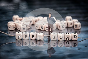 Miniature model of man and wooden cubes with feel good inscription strung on a thread on reflective table with heap of cubes