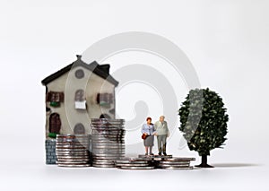 A miniature old couple on pile of coins with miniature tree and miniature building.