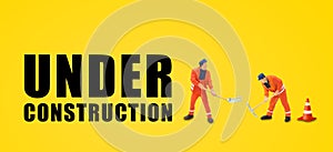 Miniature people as workers standing and working nearly traffic cone and text wordings Under Construction on yellow background.