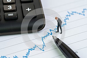 Miniature people businessman with suit standing and looking at stock price chart with calculator and pen using as financial