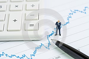 Miniature people businessman with suit standing and looking at stock price chart with calculator and pen using as financial