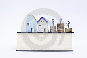 Miniature people on coins stack with wooden and ceramic vintage style house on thick book isolate on white background