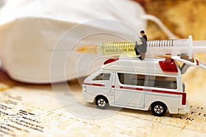 Miniature people stand on ambulance car with medical mask and Syringe of COVID-19 vaccine .
