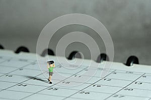 Miniature people toy figure photography. A boy running above monthly planner calendar. Grey cloudy background