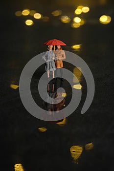 Miniature people toy figure photography. Couple dating at rainy day using umbrella at night. Beautiful golden yellow bokeh city