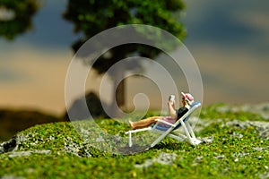 Miniature people toy figure photography. Girl seat on deck chair reading a book, relaxing at garden