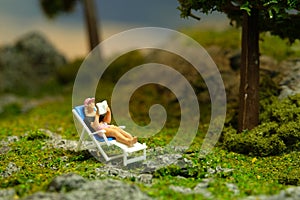 Miniature people toy figure photography. Girl seat on deck chair reading a book, relaxing at garden