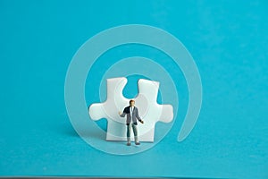 Miniature people toy figure photography. A shrugging businessman standing in front of puzzle jigsaw piece. Isolated on blue