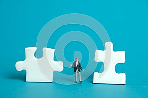 Miniature people toy figure photography. A shrugging businessman standing the middle of two puzzle jigsaw piece. Isolated on blue