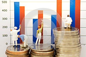 Miniature people: Worker standing on stack of coins and painting bar graph