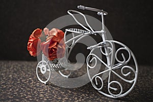 Miniature white bicycle, toy with red roses in vintage style on a brown stony background.Miniature white bicycle, toy with red