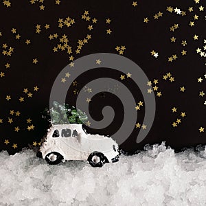 A miniature white toy car carries a Christmas tree on a roof in the snow on a dark background with shiny stars with copy space.