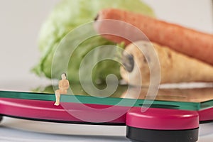 Miniature woman figure siting on the digital electronic bathroom scale for weight of human body. Fresh vegetables at shallow depth