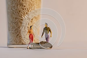 Miniature woman and man figure standing next to big glass of rice and counting coins. Shallow depth of field background. Family