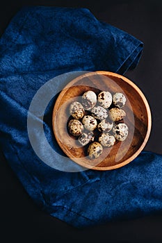 Minimal art of fresh quail eggs in the wooden bowl on the dark background with blue saten or silk around