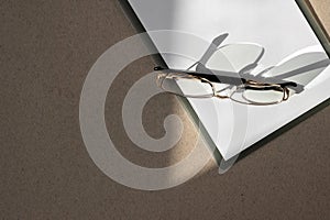 Minimal business brand template. Clipboard with white paper sheet closeup, eyeglasses on brown table with natural sunlight shadows