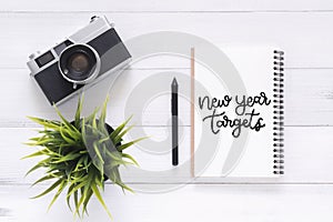 Flat lay view photo of working desk with new year target calligraphy notebook and a camera lying on white wooden background.