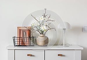 Minimalism style living room interior. Floral arrangement, table lamp, metal basket with magazines on a white chest of drawers