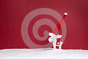 Minimalist Christmas holiday concept with white reindeer in Santa`s hat against red background with copy-space