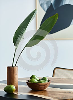 Minimalist composition on the design wooden table with fruits, tropical leaf in vase, abstract paintings and stylish chair.