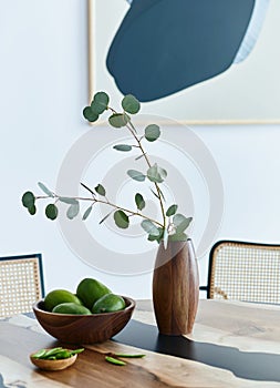 Minimalist composition on the design wooden table with fruits, tropical leaf in vase, abstract paintings and stylish chair.